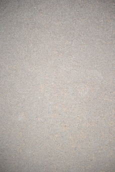 A close-up view of a textured surface that appears to be a concrete or cement wall. The surface is uniform in color with small specks and imperfections throughout, giving it a rough, gritty appearance.
