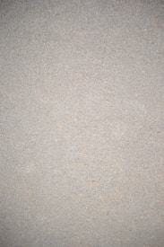 A close-up view of a textured surface that appears to be a concrete or cement wall. The surface is uniform in color with small specks and imperfections throughout, giving it a rough, gritty appearance.