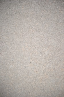 A close-up view of a textured surface that appears to be a concrete or cement wall. The surface is uniform in color with small specks and imperfections throughout, giving it a rough, gritty appearance.