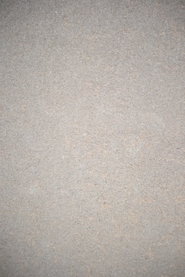 A close-up view of a textured surface that appears to be a concrete or cement wall. The surface is uniform in color with small specks and imperfections throughout, giving it a rough, gritty appearance.