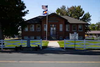 A historic brick building with a triangular roof, featuring American and Colorado flags flying outside. The building's entrance has a sign labeled 'OPEN' and is surrounded by a well-maintained lawn and white fence. Trees provide shade around the area, and a paved street runs in front of the building.
