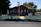 A historic brick building with a triangular roof, featuring American and Colorado flags flying outside. The building's entrance has a sign labeled 'OPEN' and is surrounded by a well-maintained lawn and white fence. Trees provide shade around the area, and a paved street runs in front of the building.