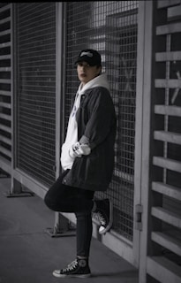 Monochrome image of a model leaning against a black metal fence, wearing layered streetwear pieces
