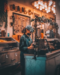 A person with curly hair wearing a face mask and a cap stands behind a counter in a warmly lit café or restaurant. They are wearing a denim apron and are focused on their tasks, surrounded by decorative elements like greenery and a chalkboard menu.