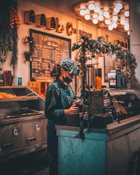 A person with curly hair wearing a face mask and a cap stands behind a counter in a warmly lit café or restaurant. They are wearing a denim apron and are focused on their tasks, surrounded by decorative elements like greenery and a chalkboard menu.