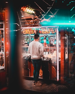 A person with short hair is standing in front of a brightly lit arcade or carnival game machine filled with plush toys. The setting features a colorful and lively atmosphere with neon lights and reflections adding to the vibrancy. The person is focused on the game controls, suggesting they are engaged in trying to win a prize.