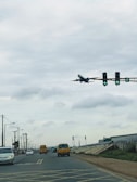 A wide urban road with several vehicles, including a white sedan and yellow buses. Overhead, an aircraft is in the midst of a low-altitude flight path against a cloudy sky. Traffic lights are visible with green arrows, and the road has visible markings. Power lines and poles line the sides of the street, with buildings and grassy areas in the background.