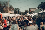 A crowded outdoor market or festival event on a sunny day, with numerous people walking between rows of white tents offering various goods. The environment includes houses in the background and large trees. The crowd is diverse, consisting of adults, teenagers, and children.