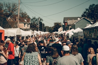 A crowded outdoor market or festival event on a sunny day, with numerous people walking between rows of white tents offering various goods. The environment includes houses in the background and large trees. The crowd is diverse, consisting of adults, teenagers, and children.