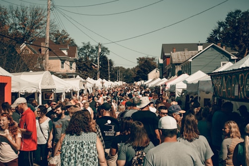 A crowded outdoor market or festival event on a sunny day, with numerous people walking between rows of white tents offering various goods. The environment includes houses in the background and large trees. The crowd is diverse, consisting of adults, teenagers, and children.