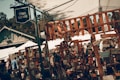 A rustic outdoor market scene with stalls displaying various handmade crafts and decorations. The area is bustling with people browsing through colorful items. Signs and decorations, such as welcome signs and wooden ornaments, are prominently displayed. A wooden fence frames much of the foreground.
