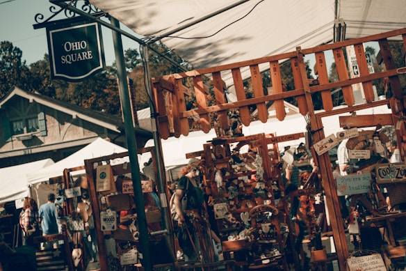 A rustic outdoor market scene with stalls displaying various handmade crafts and decorations. The area is bustling with people browsing through colorful items. Signs and decorations, such as welcome signs and wooden ornaments, are prominently displayed. A wooden fence frames much of the foreground.