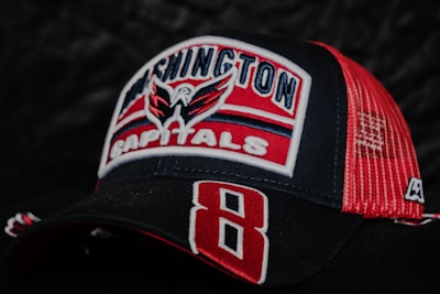 A baseball cap featuring the logo and colors of the Washington Capitals ice hockey team. The cap is predominantly black and red with the team's name and emblem embroidered on the front. The mesh back of the cap is red, and a large number 8 is stitched on the brim.