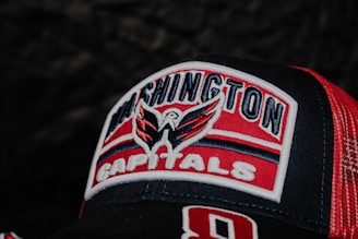 A close-up view of a Washington Capitals logo embroidered on a red and black baseball cap. The logo features a stylized eagle with spread wings above the text. The cap has a mesh pattern on the side for ventilation.