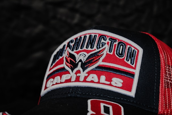 A close-up view of a Washington Capitals logo embroidered on a red and black baseball cap. The logo features a stylized eagle with spread wings above the text. The cap has a mesh pattern on the side for ventilation.
