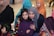 A group of people, including women and children, are gathered in what seems to be a social or family gathering. Several individuals are wearing colorful hijabs and winter clothing, suggesting a festive or warm environment.