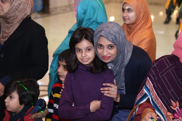 A group of people, including women and children, are gathered in what seems to be a social or family gathering. Several individuals are wearing colorful hijabs and winter clothing, suggesting a festive or warm environment.