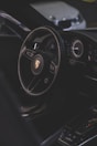 A luxury car interior focusing on the steering wheel with a recognizable logo, alongside the dashboard with modern instruments. The environment is dimly lit, providing a sleek and sophisticated ambiance.