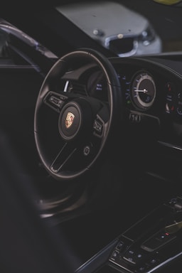 A luxury car interior focusing on the steering wheel with a recognizable logo, alongside the dashboard with modern instruments. The environment is dimly lit, providing a sleek and sophisticated ambiance.