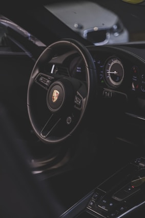 A luxury car interior focusing on the steering wheel with a recognizable logo, alongside the dashboard with modern instruments. The environment is dimly lit, providing a sleek and sophisticated ambiance.