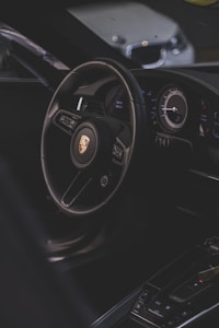 A luxury car interior focusing on the steering wheel with a recognizable logo, alongside the dashboard with modern instruments. The environment is dimly lit, providing a sleek and sophisticated ambiance.