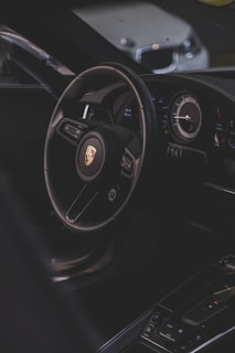 A luxury car interior focusing on the steering wheel with a recognizable logo, alongside the dashboard with modern instruments. The environment is dimly lit, providing a sleek and sophisticated ambiance.