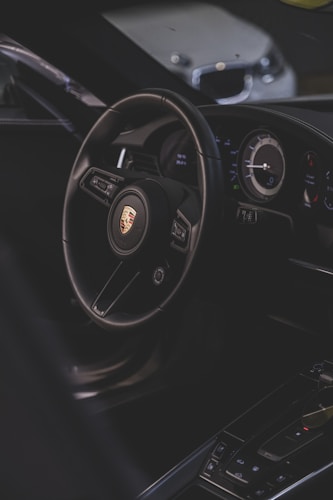 A luxury car interior focusing on the steering wheel with a recognizable logo, alongside the dashboard with modern instruments. The environment is dimly lit, providing a sleek and sophisticated ambiance.