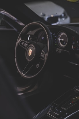 A luxury car interior focusing on the steering wheel with a recognizable logo, alongside the dashboard with modern instruments. The environment is dimly lit, providing a sleek and sophisticated ambiance.