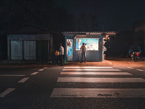 A small roadside kiosk is illuminated at night, casting a warm glow. One person stands inside the kiosk, while another stands outside holding a bag. The area is relatively dark, with the kiosk being a focal point of light. A pedestrian crossing is visible in the foreground, indicating a street setting.