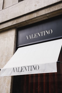 A storefront with a stone facade and a large awning displaying the word 'Valentino.' The awning is clean and white with bold black lettering, positioned above an elegant dark window.
