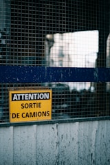 A yellow warning sign with French text reading 'ATTENTION SORTIE DE CAMIONS' is displayed on a metal mesh fence. The background is slightly blurred, possibly showing an industrial or warehouse setting.