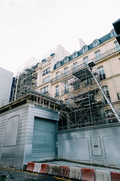 A building under renovation with extensive scaffolding surrounding its façade. The structure features classical architectural elements like ornate balconies and mansard roofs. Traffic barriers are placed at the base, indicating an active construction zone.