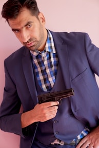 A man in a blue suit and checked shirt is holding a handgun close to his chest. He has a serious expression and is standing against a light pink background.