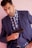 A man in a blue suit and checked shirt is holding a handgun close to his chest. He has a serious expression and is standing against a light pink background.
