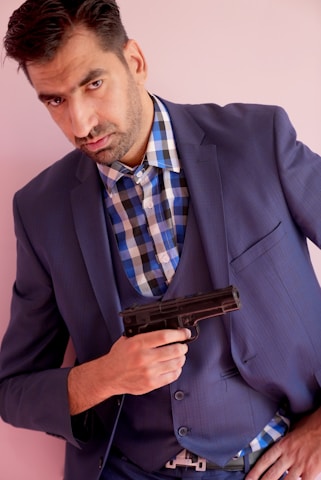 A man in a blue suit and checked shirt is holding a handgun close to his chest. He has a serious expression and is standing against a light pink background.
