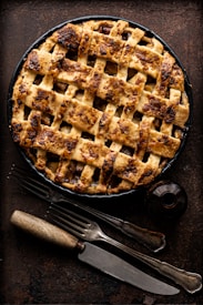 A rustic, freshly baked pie with a golden-brown lattice crust rests on a dark background. The pie is surrounded by vintage, slightly tarnished utensils including a fork and a knife with wooden handles. Nearby, there is a small, dark glass jar with a metal lid.