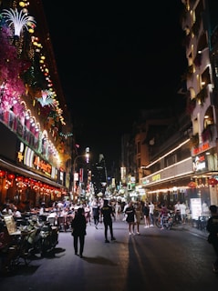 A bustling night scene on a lively city street, filled with people walking and dining outside. Bright neon lights and colorful floral decorations illuminate the surroundings, creating a vibrant atmosphere.