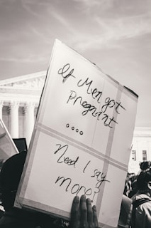 A protest sign is held up reading 'If Men got Pregnant... Need I say more?' in front of a large government building with columns, suggesting a public demonstration setting.