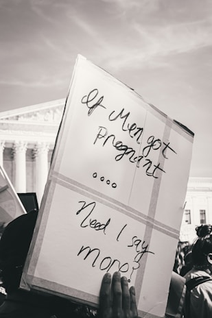 A protest sign is held up reading 'If Men got Pregnant... Need I say more?' in front of a large government building with columns, suggesting a public demonstration setting.