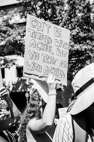 A person is holding a protest sign with the text 'Can't be trusted with a choice, but you trust me with a child?' in large, bold letters. The image is in black and white and depicts a crowd of people, suggesting a public demonstration. Trees and a building are visible in the background.