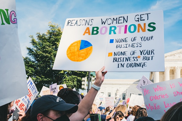 A crowd of people holding various protest signs with messages about abortion rights. The central sign features a pie chart and the message 'Reasons Women Get Abortions: None of Your Business'. The background includes trees and a government building.