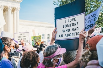A group of people is gathered outside a large, classical-style building with columns, holding signs. Some of the signs read 'I love someone who had an abortion' and 'Bans off our bodies.' Many of the individuals are wearing masks and hats, suggesting a focus on personal safety and the gravity of the cause.