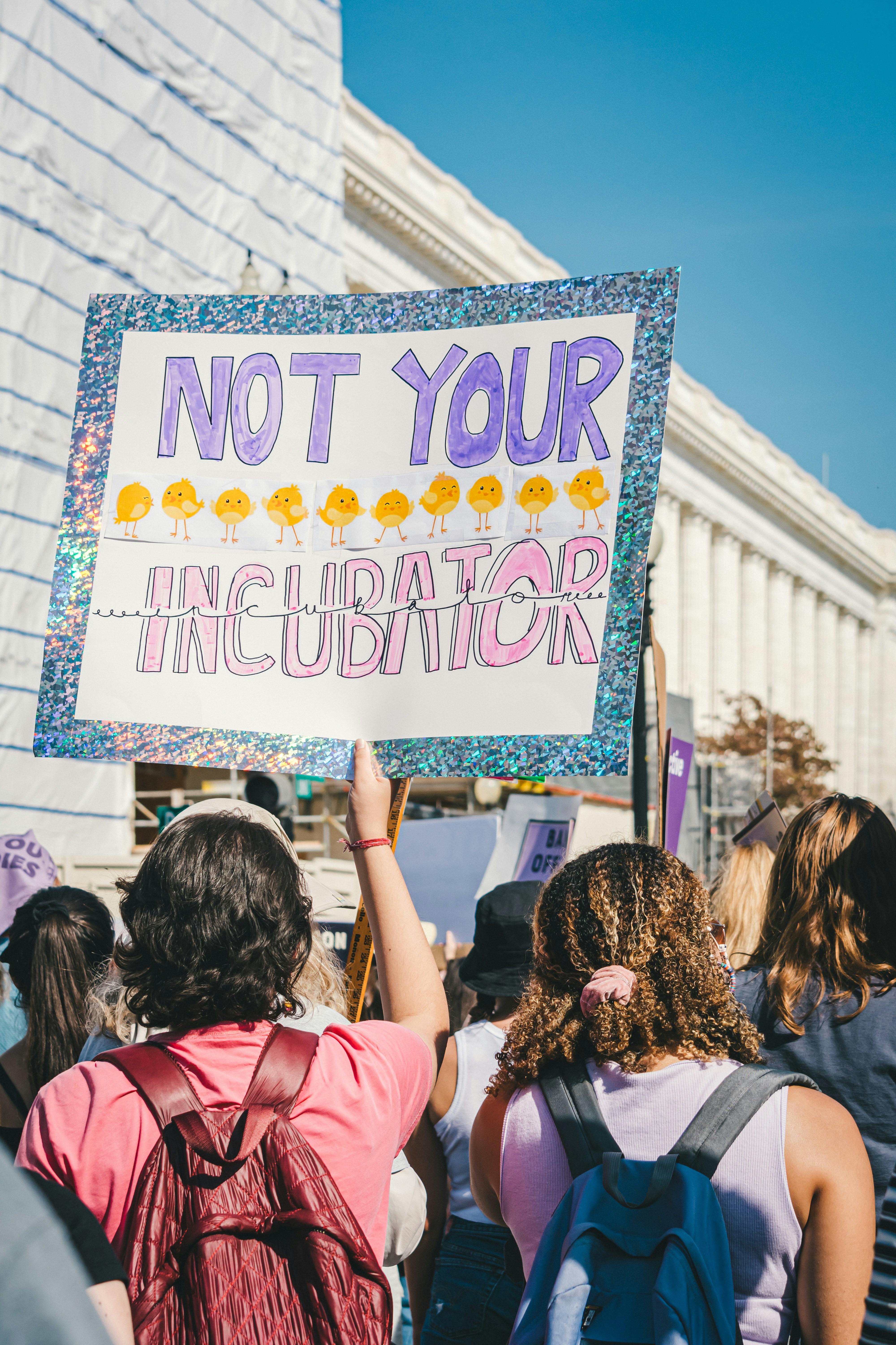 In a crowd of protestors, one person holds up a sign that says "Not Your Incubator" in purple and pink marker with hand-drawn yellow chicks.