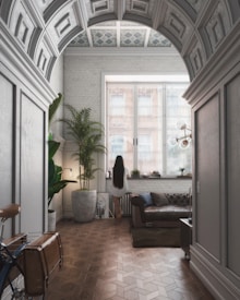 A stylish interior room with a vaulted ceiling and intricate patterns. Natural light streams through large windows, illuminating the space. A person with long hair stands by the window, surrounded by modern furniture such as a leather sofa and large potted plants. A bicycle is partially visible in the foreground, adding an eclectic touch.