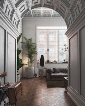 A stylish interior room with a vaulted ceiling and intricate patterns. Natural light streams through large windows, illuminating the space. A person with long hair stands by the window, surrounded by modern furniture such as a leather sofa and large potted plants. A bicycle is partially visible in the foreground, adding an eclectic touch.