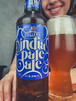 A stylish bottle of Fuller's India Pale Ale with a vibrant blue label featuring ornate lettering sits next to a full glass of amber-colored beer. A person smiling in the background adds a cheerful atmosphere.