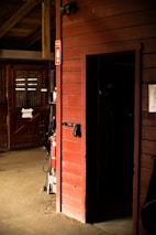 An indoor stable area with wooden paneling and a door leading to a dark room. There are numbered stalls, one of which is seen with a half-door and a window behind metal bars. A fire extinguisher is mounted on the wall with a visible 'Fire' sign. Various objects such as a saddle, bridle, and a tack are hanging near the stall. The floor is made of dirt, adding to the rustic atmosphere.