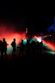 Silhouettes of people standing in a dark space are illuminated by colorful lights. Red and green lights create a dramatic, atmospheric effect with fog or smoke dispersing the beams.