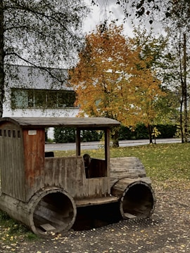 A rustic wooden play structure designed to resemble a vehicle sits on a grassy area. The structure is made from large wooden logs, with a steering wheel visible. In the background, autumn-colored trees with golden leaves stand against a cloudy sky. A modern building partially visible through the tree branches contrasts with the natural setting.