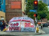A street corner with an urban setting, featuring a large tent with bold graffiti-style writing advocating social issues. Behind the tent is a brick building and a traffic light showing red. There are several people standing nearby, and a bus is visible in the background.